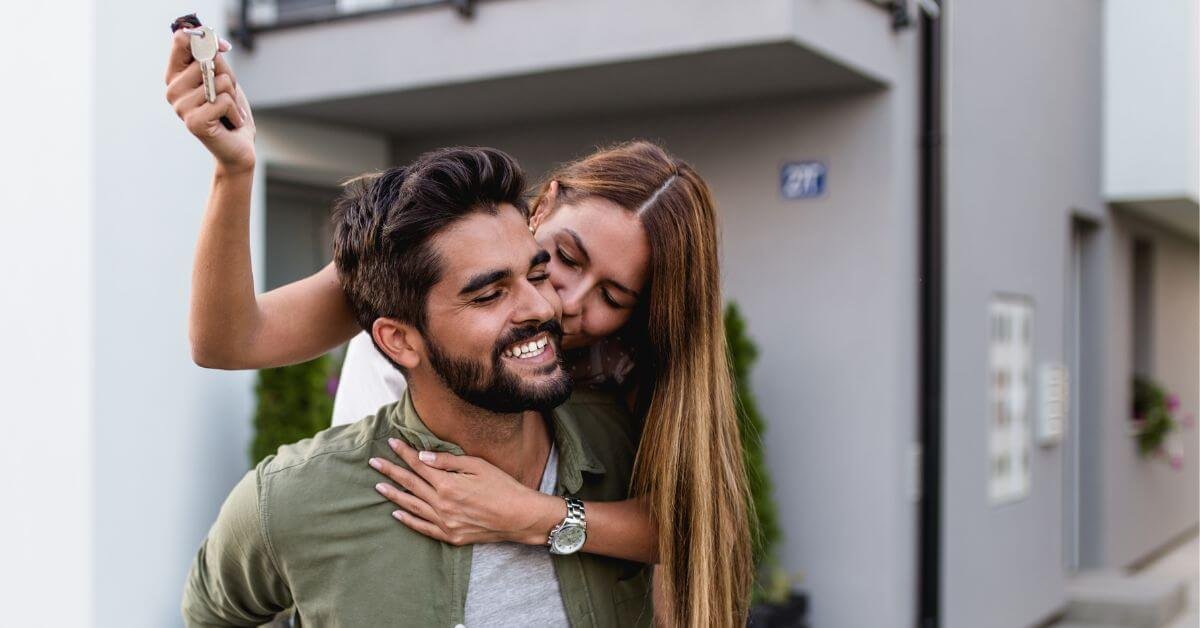 Photo of a happy couple outside of the new home that they just bought. The man is smiling as the woman embraces him from behind