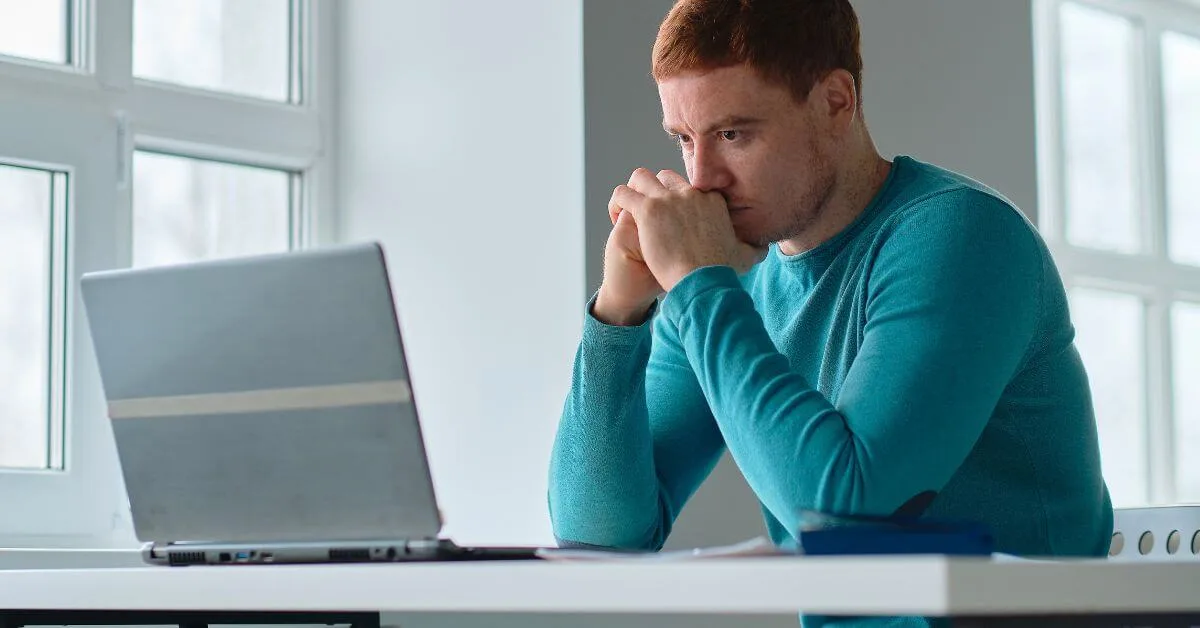 Concentrated man in teal sweater working on laptop in a bright room.