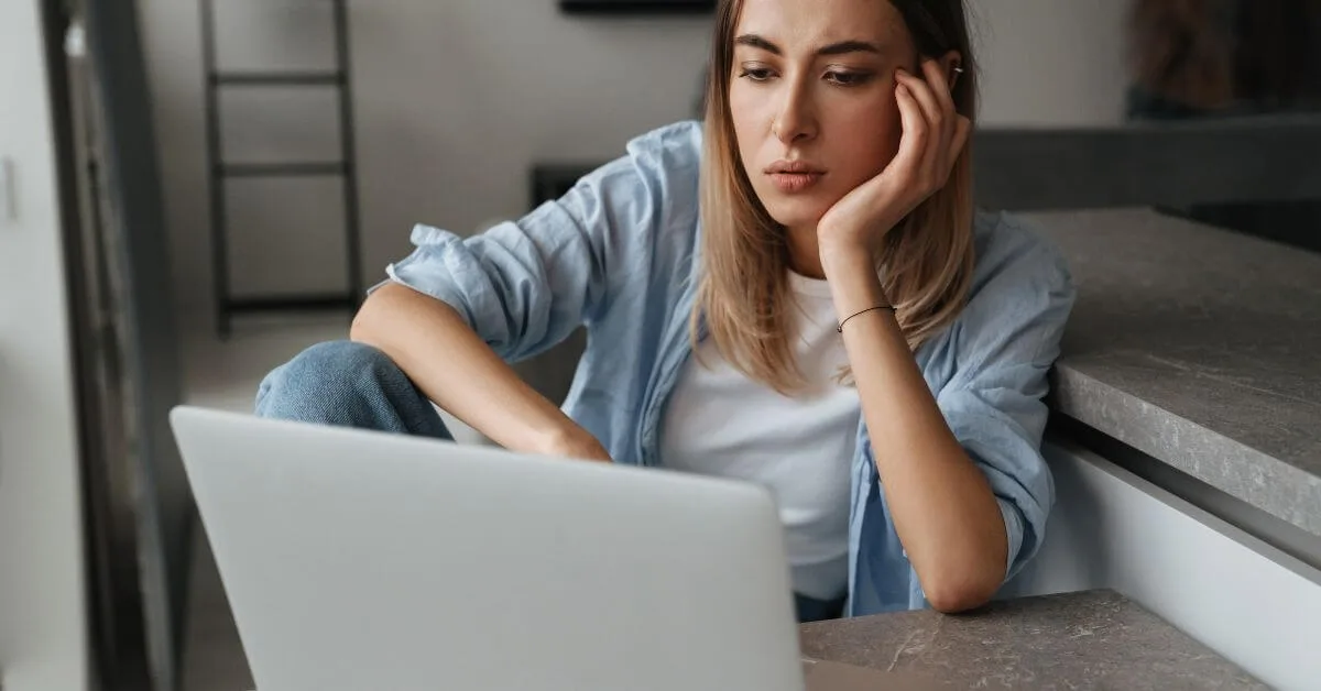 A young woman looking at her laptop with a contemplative expression.