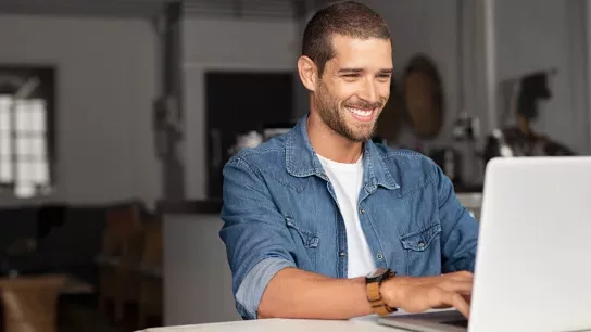 Cheerful man in denim shirt using a laptop at home.