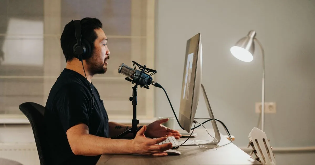 Man with headphones speaking into a microphone in front of a computer monitor, desk lamp on the side