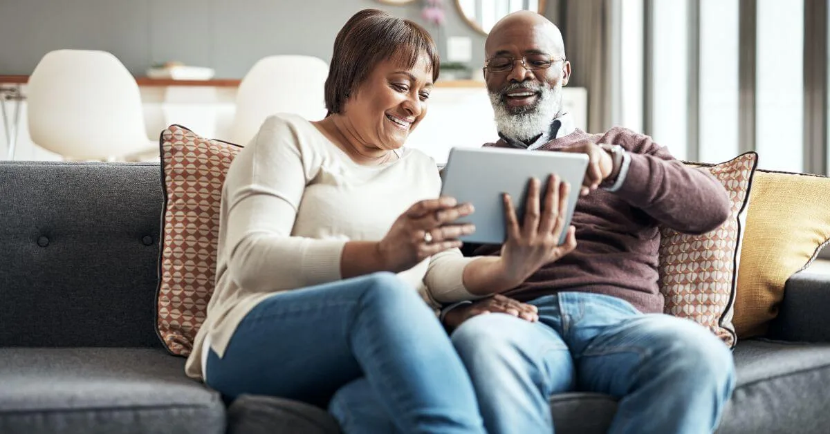 An older couple smiling while looking at a tablet together on their living room couch.