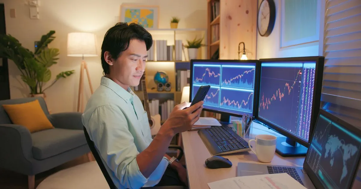 Man at home office desk using smartphone and computer screens to monitor his investments.